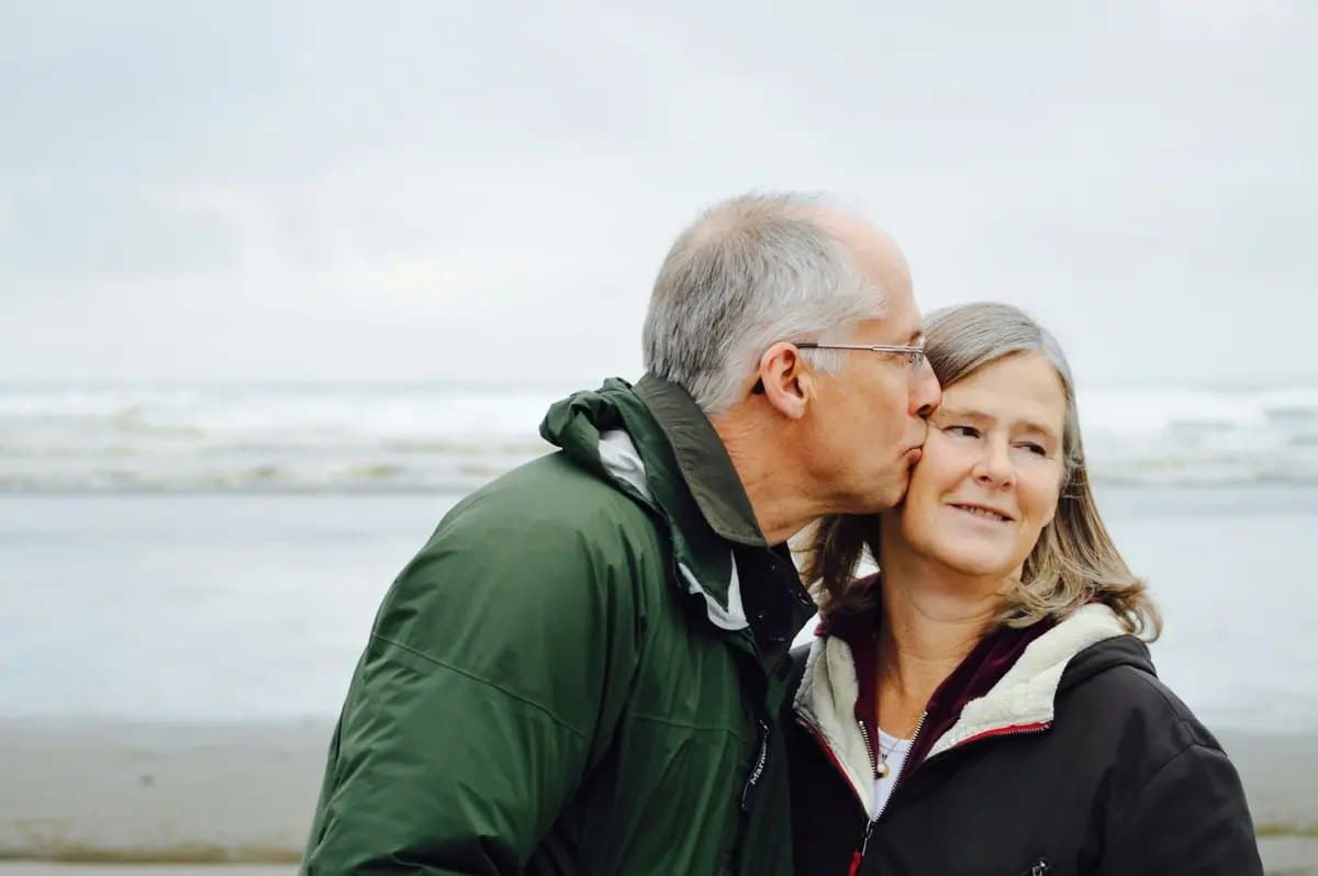 An older couple on a windswept beach; he kisses her on the cheek and she smiles