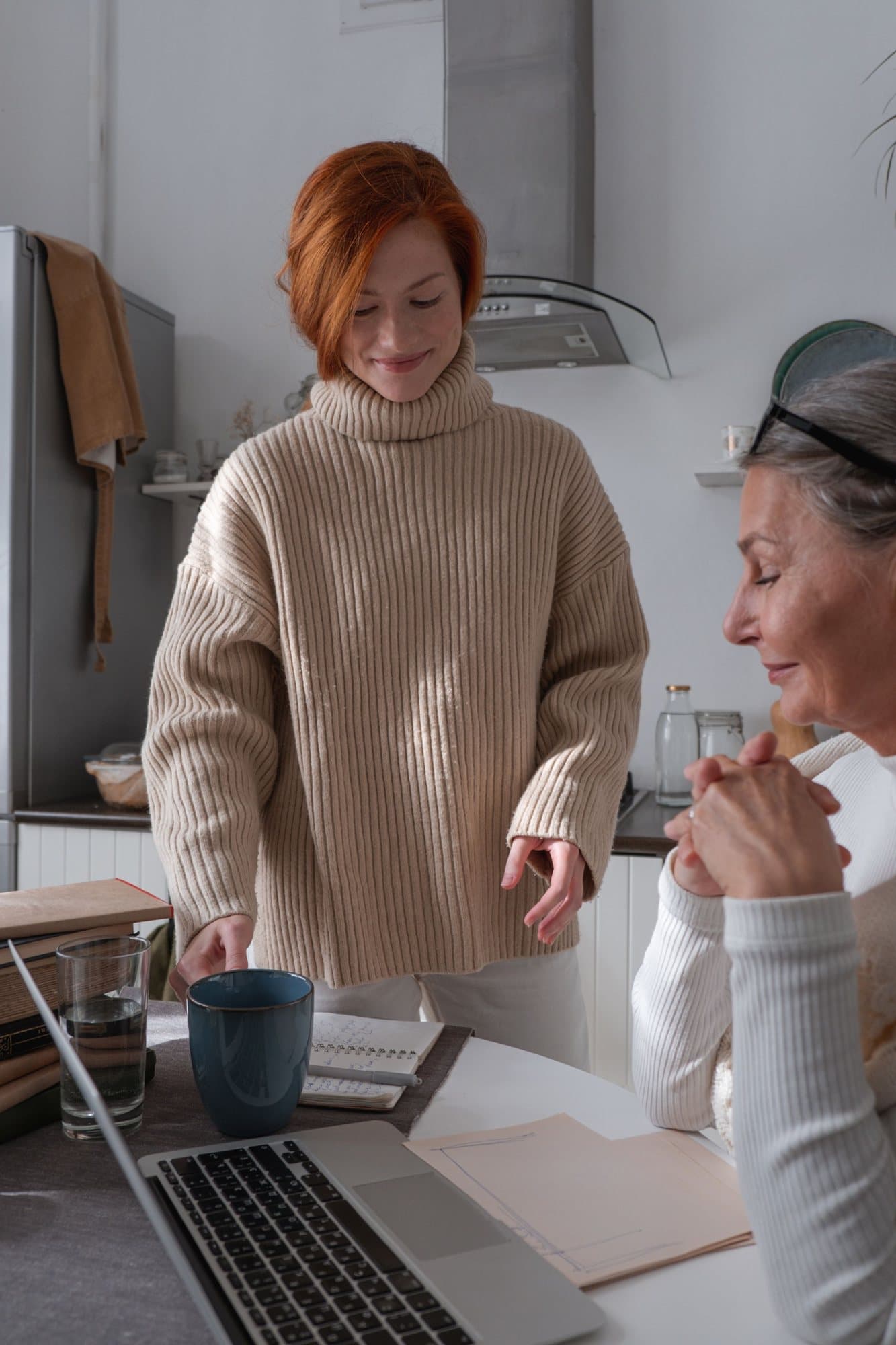 An older woman and a younger woman at a kitchen table, looking at a laptop together